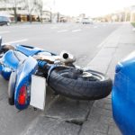 The accident blue bike with a blue car. The motorcycle crashed into the bumper of the car on the road. The motorcycle lies on the road near the car.