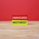two wooden blocks labeled Uninsured and Motorist, stacked on a wooden table against an red background. The image conveys insurance gaps, risk on the road, and driver protection.