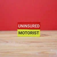 two wooden blocks labeled Uninsured and Motorist, stacked on a wooden table against an red background. The image conveys insurance gaps, risk on the road, and driver protection.